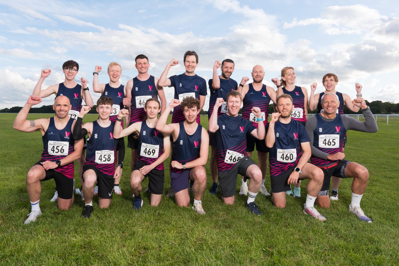 Fifteen Vale of York Athletic Community runners in navy and pink club vests, arms raised and cheering, at the York and District Road Race League on the Knavesmire